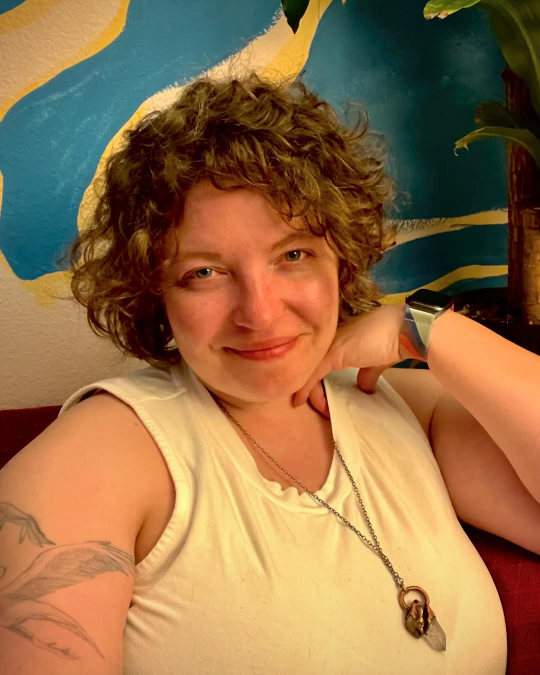 A woman with curly brown hair wearing a cream-colored sleeveless top and a bronze & copper pendant necklace, posing in front of a blue and yellow painted wall.