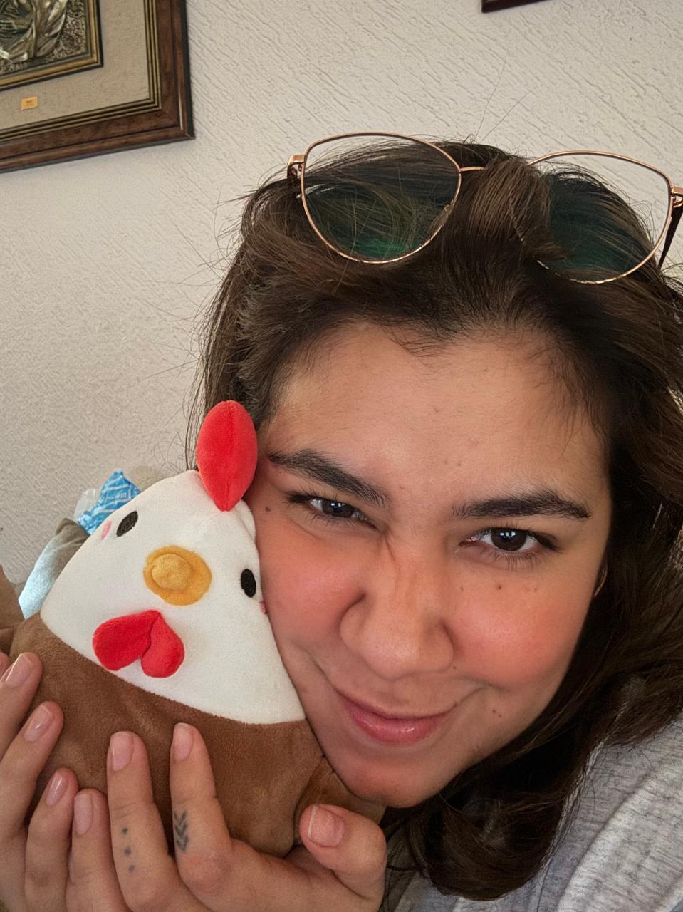 A close-up selfie of a woman with wire rimmed glasses on her head, holding a plush toy chicken next to her face. The plush chicken has a white head, red comb and wattle, a yellow beak, and a brown body.