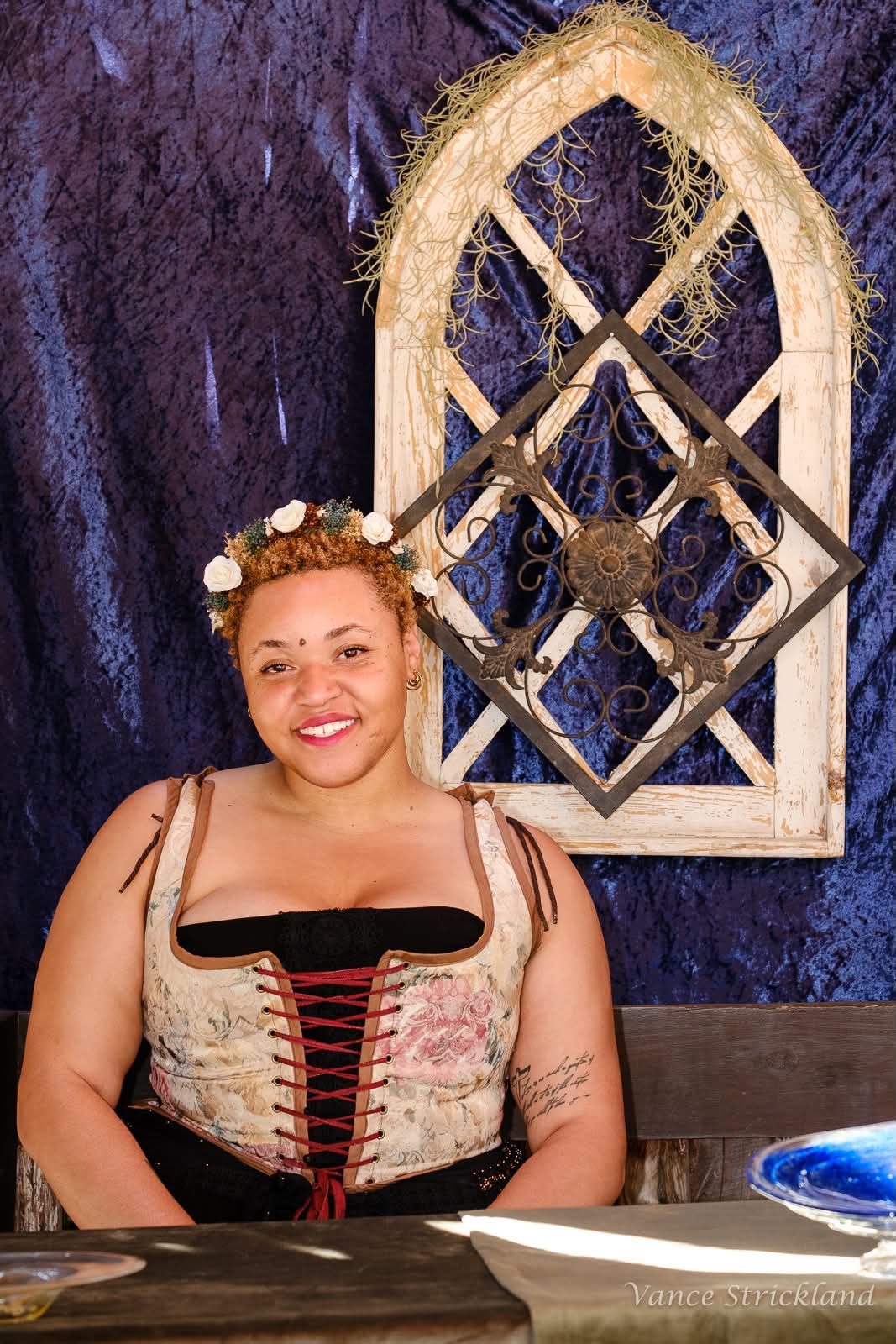 A woman wearing a vintage-style floral corset top with red lacing sits in front of a dark blue textured backdrop and a rustic wooden arched wall decoration.