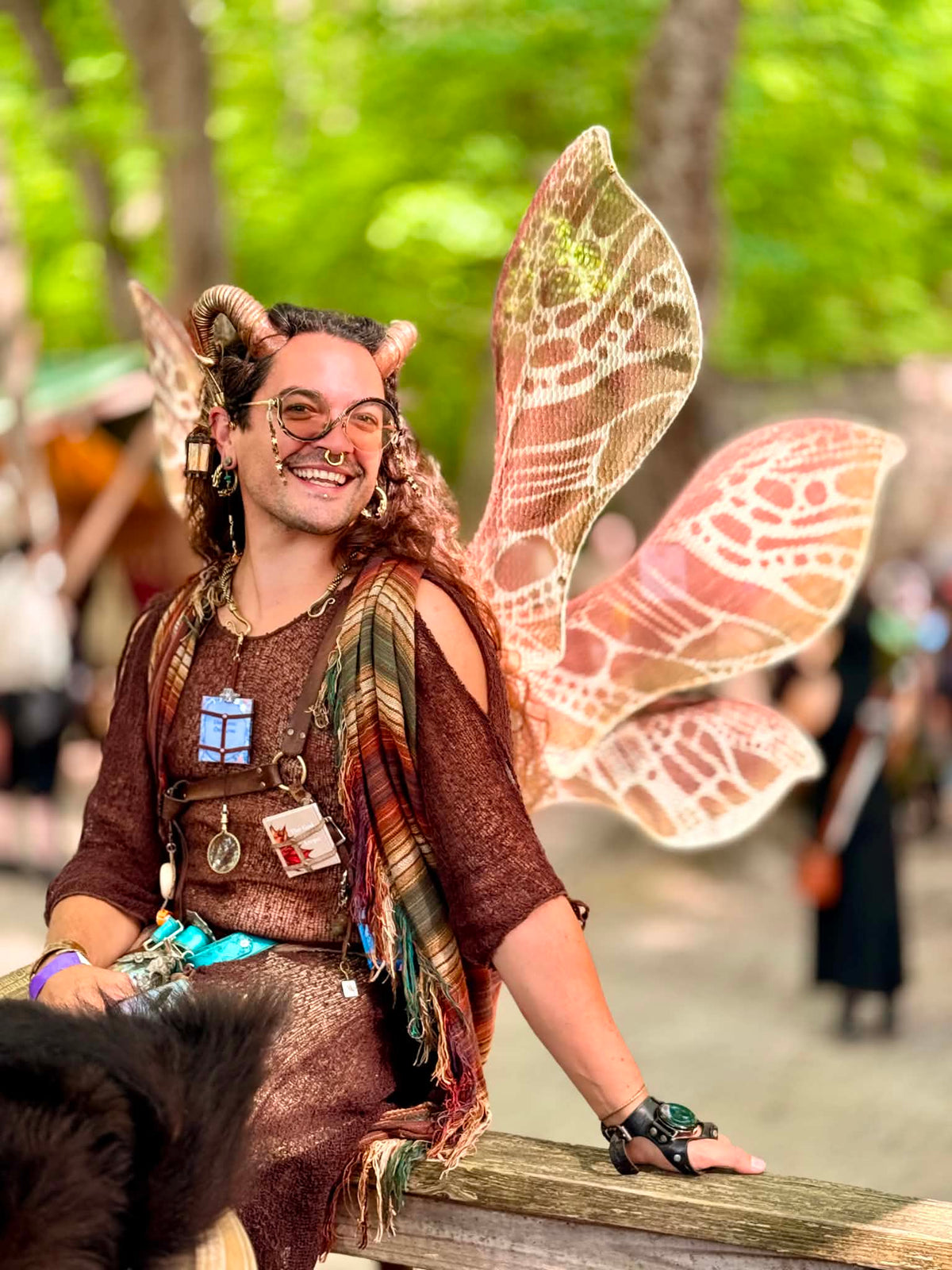A person outdoors wearing a fantasy costume with large patterned fairy wings, a brown mesh top and skirt, a patterned shawl, and horn headpiece, photographed in a festival setting.