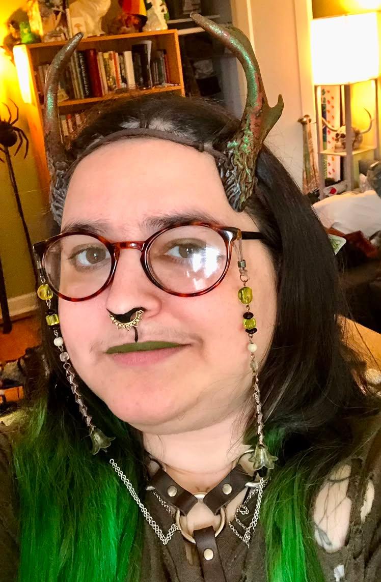 A person wearing a horned headpiece with green and bronze tones, round red-rimmed glasses, and long dangling beaded spectacle-chains, photographed indoors in a room with bookshelves and warm lighting.