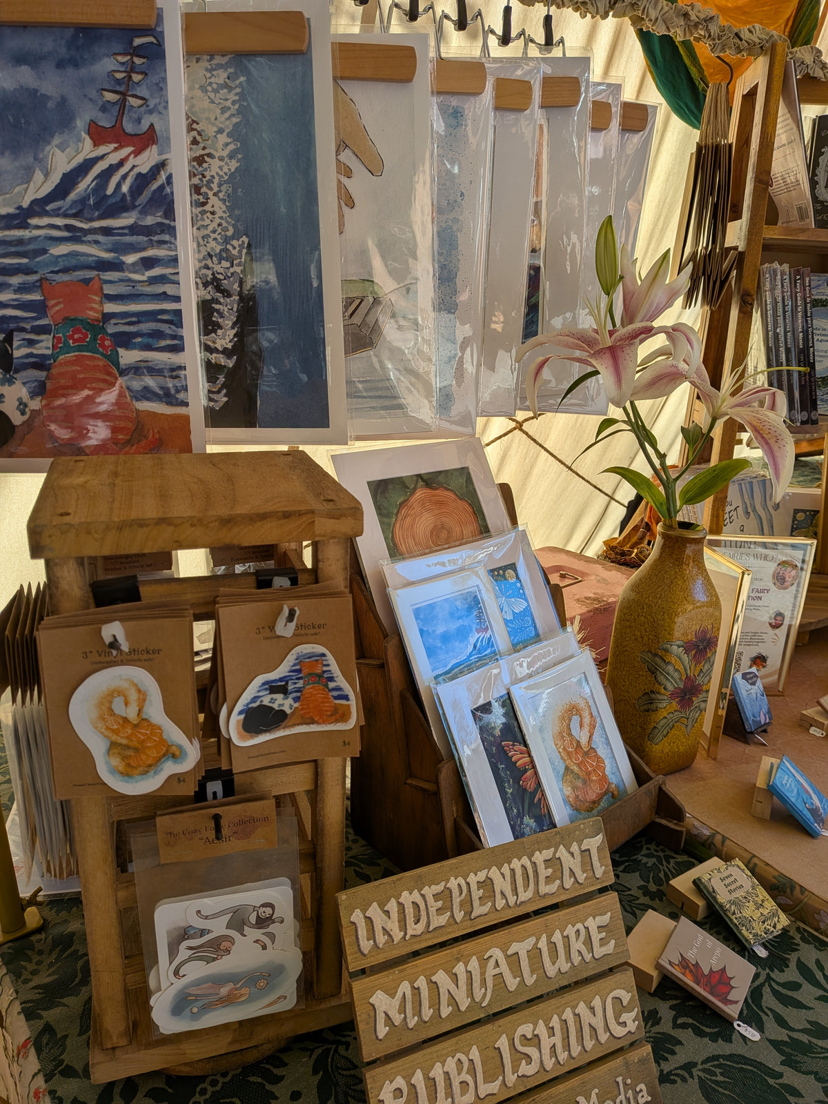 A display of handmade miniature art prints, stickers, and cards arranged on a table inside a craft market stall. Visible items include small illustrated cards, a wooden display stand with die-cut stickers, a vase with pink lilies, and a sign reading 'INDEPENDENT MINIATURE PUBLISHING'.